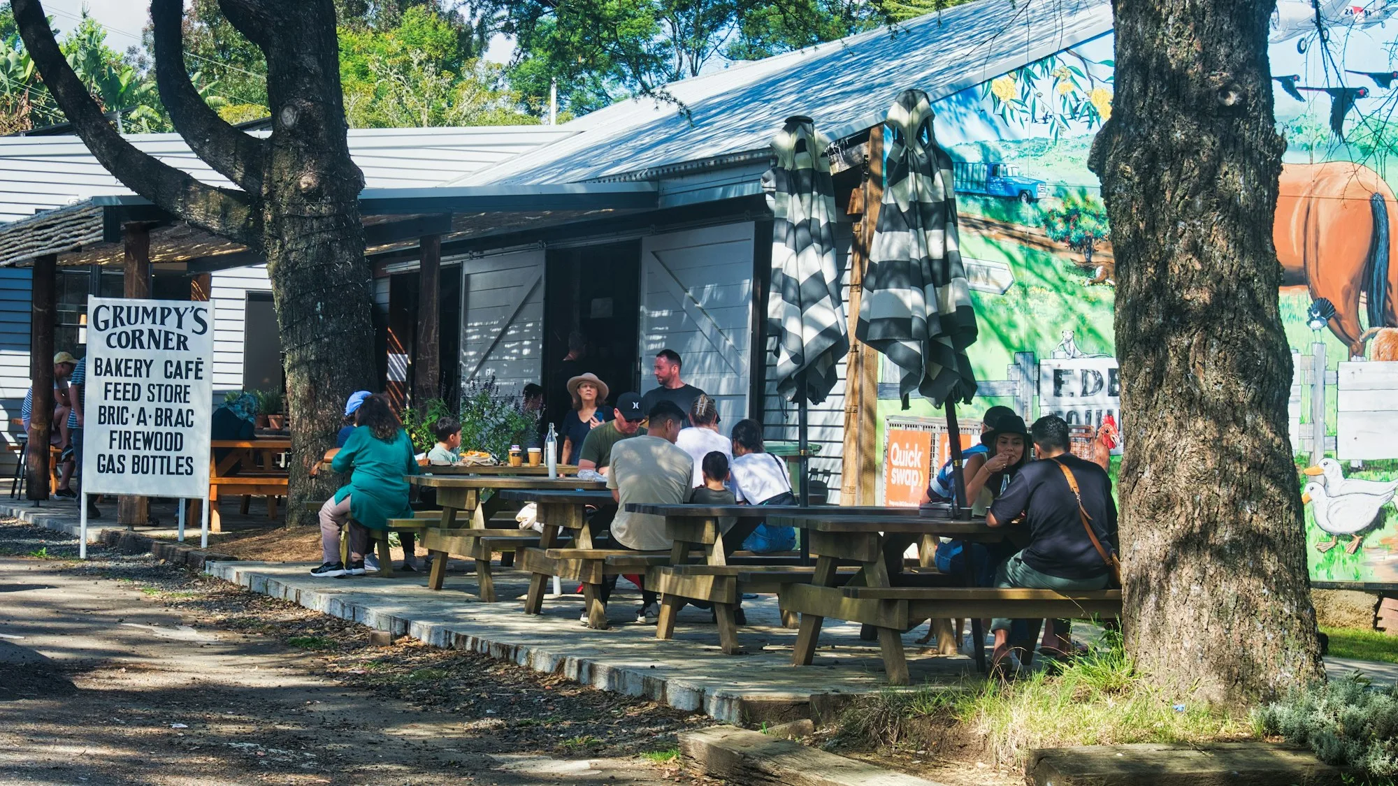 People sharing food at outdoor timber tables beside a country bakery café, dappled shade from large trees