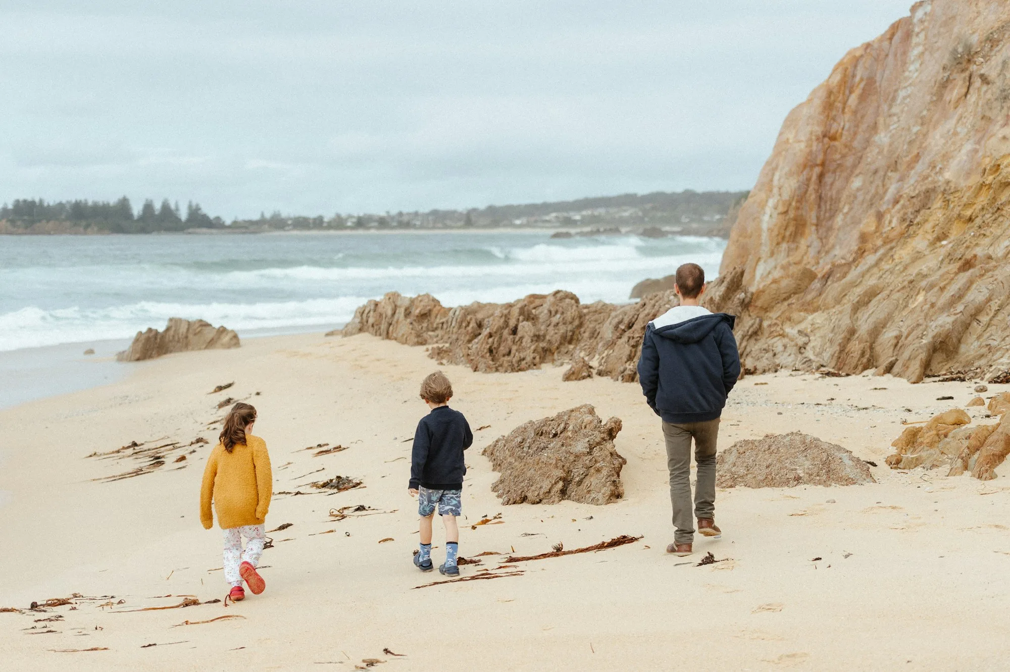 An adult and two children, seen from behind, walking along a sandy beach beside ochre rocks