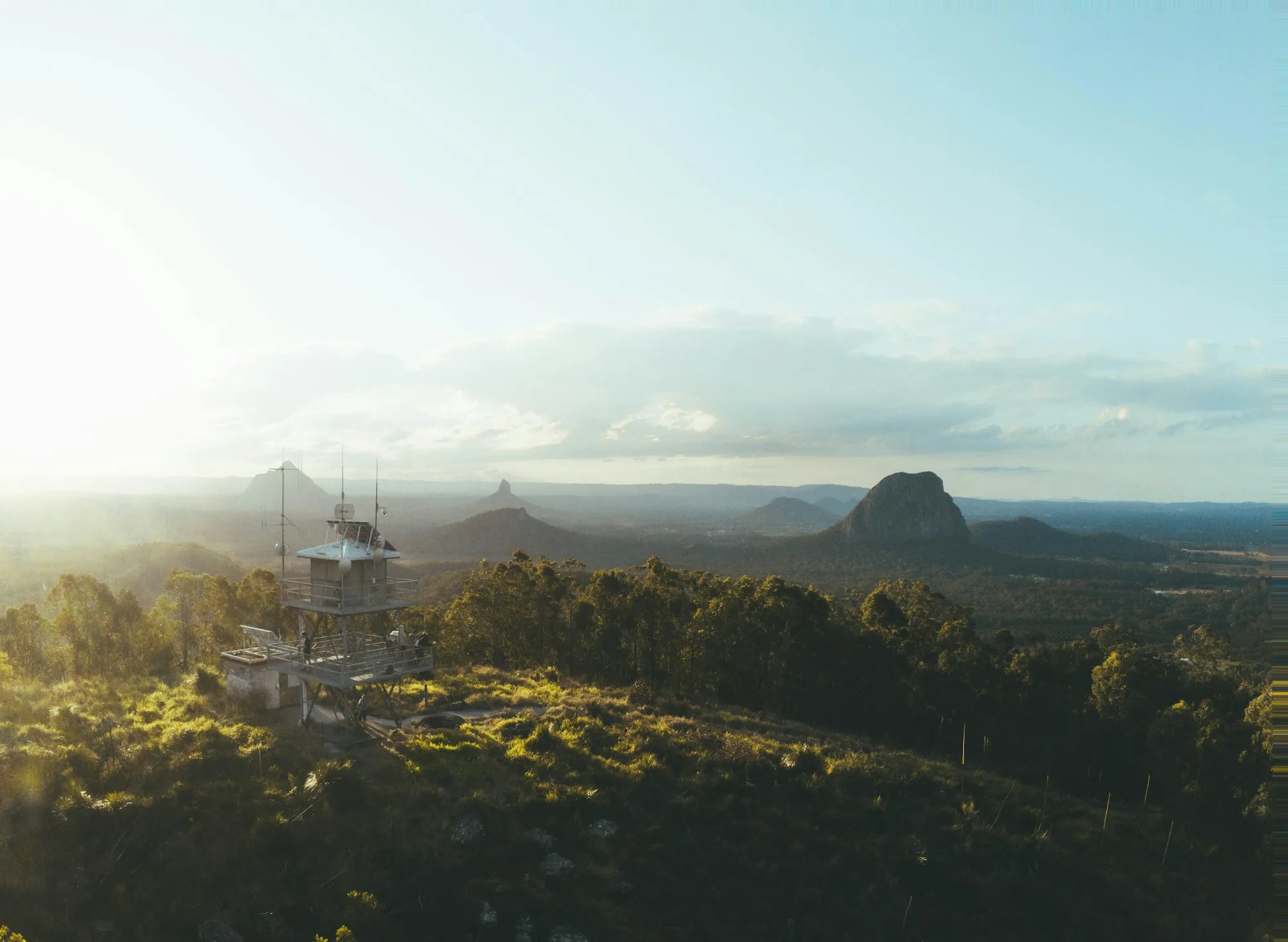 Late sun over forested hills and the volcanic peaks of the Glass House Mountains, with a lookout tower in the foreground